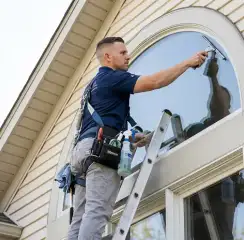 Professional window cleaner on a ladder using a squeegee to clean an exterior arched window, Knoxville, TN.
